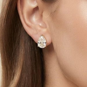 May include: A pear-shaped stud earring featuring a clear, faceted gemstone set in a metallic prong setting. The earring is positioned on the earlobe, showcasing the teardrop silhouette and the reflective, brilliant cut of the stone.