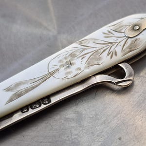 May include: A folding pocket knife featuring a smooth white handle with an intricate engraved floral pattern. The metal bolster and frame display visible hallmarks. A curved metal hook extends from the side, and a metal loop is attached to the end.