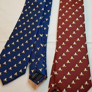 May include: Two neckties, one blue and one maroon, both featuring a repeating pattern of small penguins. Each tie has a black label on the back indicating 100% polyester composition. The ties are displayed side by side on a light surface.