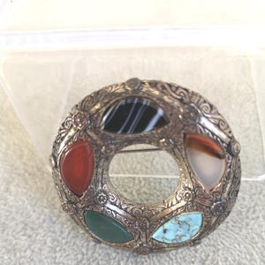 May include: A circular silver-toned metal brooch featuring an intricate embossed pattern and four teardrop-shaped inlaid stones. The stones include a black and white striped agate, a translucent brown agate, a solid green stone, and a blue turquoise.