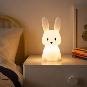 May include: A white, rabbit-shaped LED lamp that emits a soft, warm glow. The lamp features long, upright ears, two black oval eyes, and a small black nose. It is constructed from a smooth, matte material and sits upright on a flat surface.