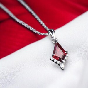 May include: A silver-toned necklace featuring a kite-shaped pendant with a deep red gemstone at its center. The pendant is accented with a series of small, clear, round-cut stones along the lower edges and a single pear-shaped stone at the base.