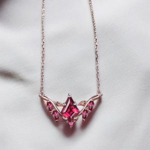 May include: A rose gold necklace featuring a central kite-shaped deep red gemstone flanked by smaller marquise-cut red stones arranged in a leaf-like pattern. The delicate metal chain connects to the ornate setting, creating a symmetrical design.
