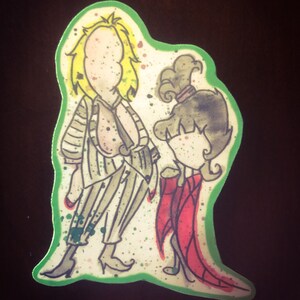 May include: A green and white sticker with a cartoon illustration of a man and a woman. The man is wearing a striped shirt and pants and has blonde hair. The woman is wearing a red dress and has brown hair. The sticker has a green border.