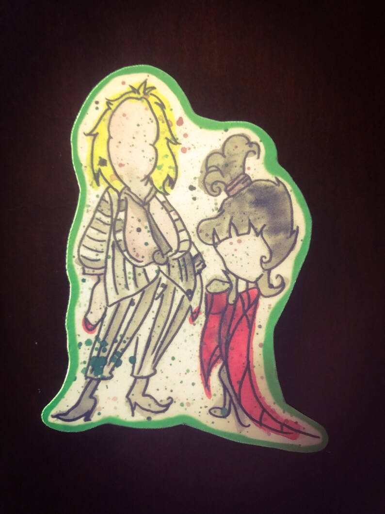 May include: A green and white sticker with a cartoon illustration of a man and a woman. The man is wearing a striped shirt and pants and has blonde hair. The woman is wearing a red dress and has brown hair. The sticker has a green border.
