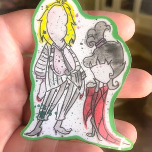 May include: A sticker featuring a cartoon illustration of two characters. The character on the left is wearing a striped suit and has yellow hair. The character on the right is wearing a red dress and has black hair. The sticker has a green border and a white background.