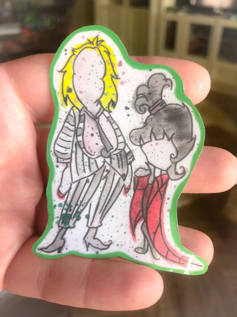 May include: A sticker featuring a cartoon illustration of two characters. The character on the left is wearing a striped suit and has yellow hair. The character on the right is wearing a red dress and has black hair. The sticker has a green border and a white background.