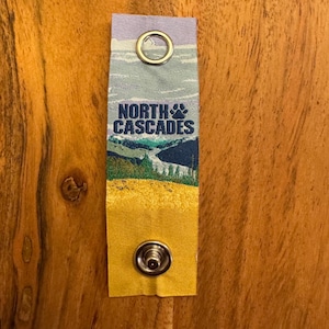 May include: A rectangular fabric strap featuring a landscape print of the North Cascades with a mountain range, forest, and yellow field. The text NORTH CASCADES with a paw print icon is centered. It includes a silver metal grommet and a snap fastener.