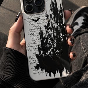 May include: A white smartphone case featuring a black silhouette of a gothic castle and a small bat icon. The background is covered in black cursive handwriting text. The case has a cutout for a triple-lens camera system on the top left corner.