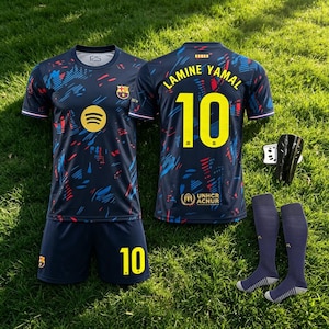 Puede incluir: Un conjunto de fútbol azul oscuro que incluye una camiseta con un patrón abstracto rojo y azul, el escudo del FC Barcelona, un logo de Spotify amarillo y pantalones cortos a juego con el número 10. La espalda muestra LAMINE YAMAL 10, espinilleras y calcetines.