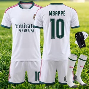 以下が含まれることがあります： ダークグリーンとピンクのアクセントが入った、白のサッカーユニフォームセット。シャツには「Emirates FLY BETTER」の文字、レアル・マドリードのエンブレム、MBAPPÉの名前と背番号10がプリントされています。すね当てとソックスが含まれています。