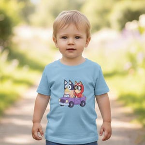 Pode incluir: Uma t-shirt de manga curta azul-clara com uma estampa gr&aacute;fica de dois c&atilde;es de desenho animado num carro roxo. Um c&atilde;o &eacute; azul com &oacute;culos pretos e o outro veste uma camisola vermelha com capuz. Gola redonda e bainha reta.