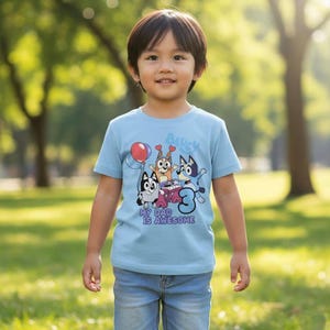 Pode incluir: Uma t-shirt de manga curta azul-clara com um gr&aacute;fico de tr&ecirc;s c&atilde;es de desenho animado, um bal&atilde;o vermelho, um teclado cor-de-rosa e o n&uacute;mero tr&ecirc;s. O texto "Bluey" e "My Dad Is Awesome" est&aacute; impresso em azul e roxo na frente.