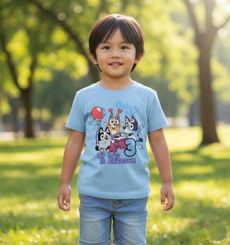 Pode incluir: Uma t-shirt de manga curta azul-clara com um gr&aacute;fico de tr&ecirc;s c&atilde;es de desenho animado, um bal&atilde;o vermelho, um teclado cor-de-rosa e o n&uacute;mero tr&ecirc;s. O texto "Bluey" e "My Dad Is Awesome" est&aacute; impresso em azul e roxo na frente.