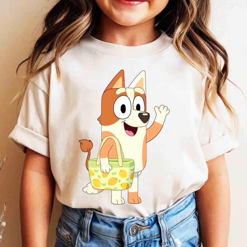 Pode incluir: Uma t-shirt de manga curta cor de creme com o gr&aacute;fico de um c&atilde;o de desenho animado com pelo laranja e branco. O c&atilde;o sorri, acena com uma pata e carrega um saco de compras verde claro decorado com um padr&atilde;o de lim&otilde;es amarelos.