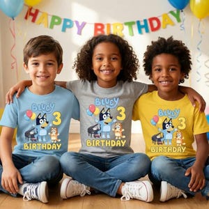 Pode incluir: Tr&ecirc;s t-shirts em azul claro, cinzento e amarelo, cada uma com um gr&aacute;fico das personagens de desenhos animados Bluey e Bingo, um piano, bal&otilde;es e o texto Bluey 3 Birthday. As t-shirts s&atilde;o usadas por tr&ecirc;s crian&ccedil;as sentadas em fila.