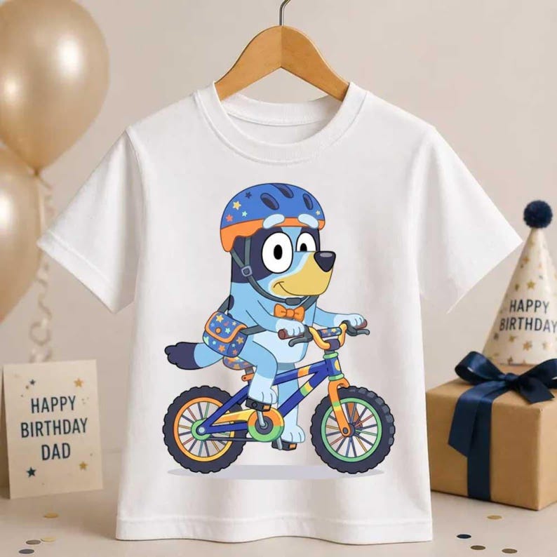 Pode incluir: Uma t-shirt branca de manga curta com o gr&aacute;fico de um c&atilde;o azul de desenho animado a usar um capacete laranja e uma gravata borboleta laranja, a andar de bicicleta. O c&atilde;o tem uma mala estampada e est&aacute; centrado na pe&ccedil;a.