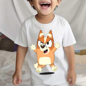 Pode incluir: Uma t-shirt branca de manga curta com uma impress&atilde;o gr&aacute;fica de um c&atilde;o de desenho animado cor de laranja e creme com orelhas pontiagudas, uma express&atilde;o de piscar de olhos e bra&ccedil;os levantados, centrada na frente da pe&ccedil;a de gola redonda.