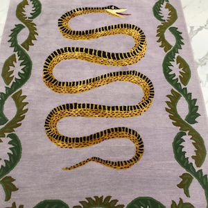 May include: A rectangular rug featuring a light lavender background with a central yellow and black patterned snake motif. The border consists of a repeating dark green leafy vine pattern that frames the snake on both the left and right sides.