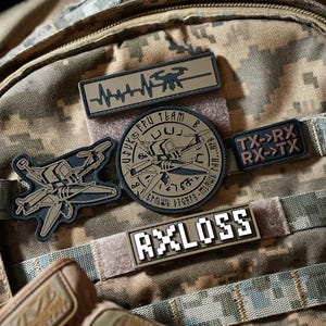 Könnte beinhalten: Ein Tarnrucksack mit mehreren taktischen Aufnähern. Die Aufnäher zeigen eine Drohnen-Herzschlaglinie, ein kreisförmiges FPU-Team-Drohnenemblem, einen TX-RX-Signalaufnäher und einen rechteckigen Aufnäher mit dem verpixelten Text RXLOSS.