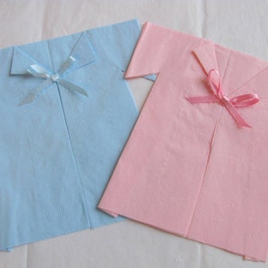 May include: Two folded paper napkins shaped like shirts, one light blue with a light blue ribbon and one pink with a pink ribbon.