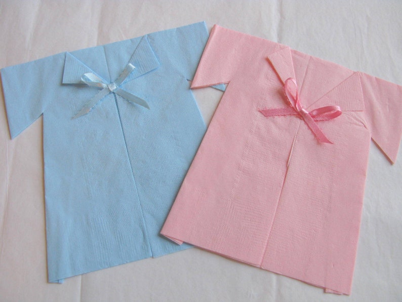 May include: Two folded paper napkins shaped like shirts, one light blue with a light blue ribbon and one pink with a pink ribbon.