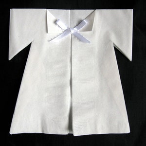 May include: A white paper napkin folded into the shape of a baby's onesie with a white ribbon tied at the neck.