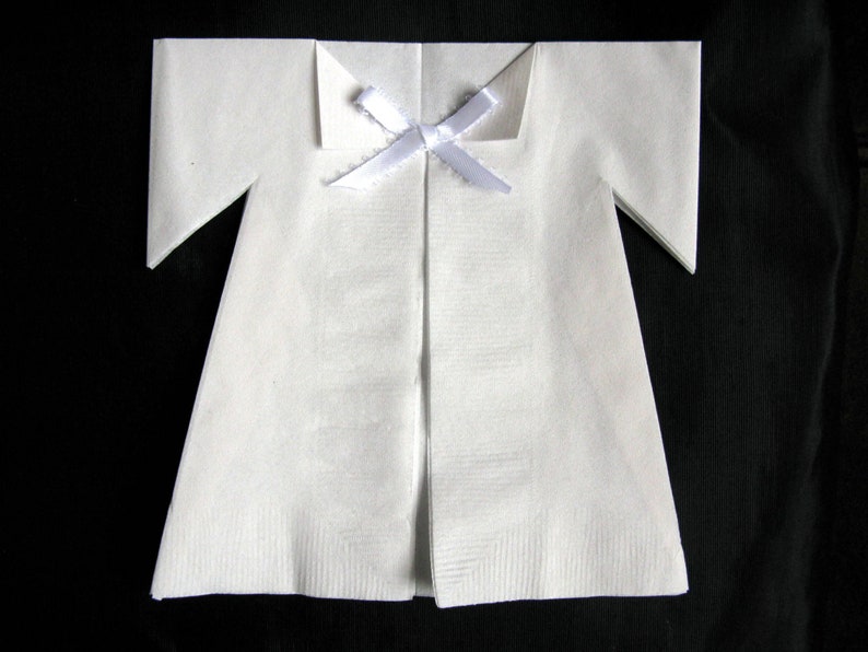 May include: A white paper napkin folded into the shape of a baby's onesie with a white ribbon tied at the neck.