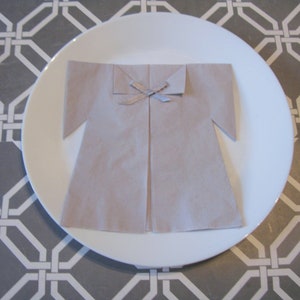 May include: A beige napkin folded into the shape of a dress with a silver ribbon tied at the neck. The napkin is on a white plate with a gray and white geometric pattern background.