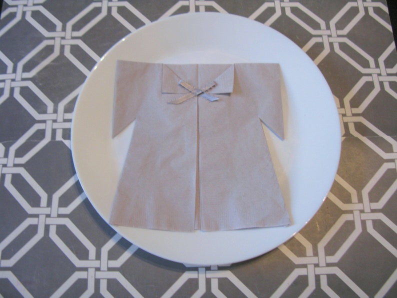 May include: A beige napkin folded into the shape of a dress with a silver ribbon tied at the neck. The napkin is on a white plate with a gray and white geometric pattern background.