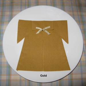 May include: A gold napkin folded into the shape of a dress with a gold ribbon tied around the neck. The napkin is on a white plate with a plaid tablecloth.