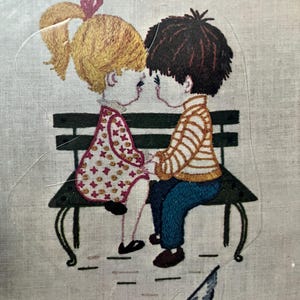 May include: Embroidered image of two children sitting on a park bench, holding hands. The girl has blonde hair in a ponytail with a red bow, and the boy has dark hair. They are wearing colorful clothing. A bird is pecking at the ground.