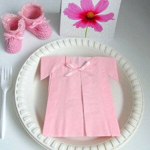 May include: A pink napkin folded into the shape of a baby dress with a pink ribbon bow. The dress is on a white plate with a white fork to the left. There are two pink crocheted baby booties in the upper left corner of the image.