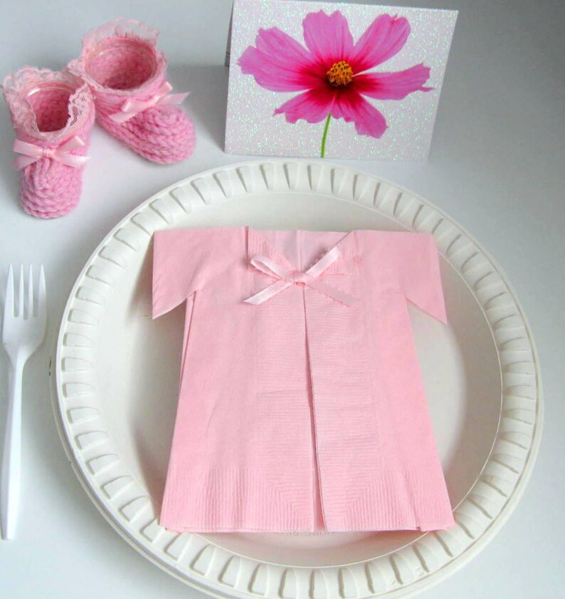 May include: A pink napkin folded into the shape of a baby dress with a pink ribbon bow. The dress is on a white plate with a white fork to the left. There are two pink crocheted baby booties in the upper left corner of the image.