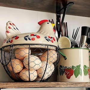 May include: A cream-colored ceramic chicken-shaped egg basket with a black wire mesh base, decorated with red and blue floral patterns. The basket is filled with brown eggs and sits next to a cream-colored metal utensil holder with strawberry motifs.