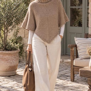 Peut inclure: Un poncho en tricot beige avec un col roulé côtelé et un ourlet pointu, porté sur un haut blanc à manches longues et un pantalon blanc à jambes larges, accompagné d'un sac fourre-tout en cuir marron et de baskets blanches à semelles fauves.