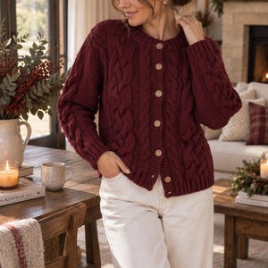 May include: A deep burgundy cable-knit cardigan featuring a round neckline, long sleeves with ribbed cuffs, and a front button closure with six light-colored wooden buttons. The knit pattern includes vertical cable textures and ribbed hem.