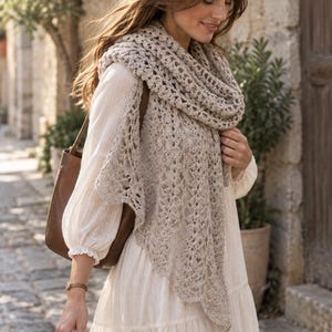 May include: A beige crochet shawl with an intricate openwork pattern draped over a long-sleeved white tiered dress, paired with a brown leather shoulder bag. The shawl features a scalloped edge and a textured, lightweight yarn construction.