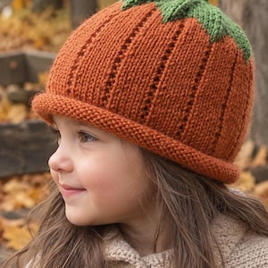 May include: A knitted beanie hat in a vibrant pumpkin orange color, featuring a textured ribbed pattern with vertical eyelet details. The top of the hat is adorned with a green knitted stem and leaf design, and it has a rolled brim at the base.