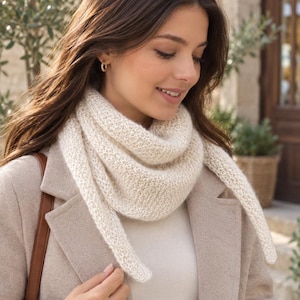 Peut inclure: Une écharpe triangulaire en tricot de couleur crème avec un motif texturé à mailles ouvertes. L'écharpe est drapée autour du cou, avec des extrémités pointues qui tombent. Elle est portée sur un manteau beige clair et un haut assorti.