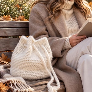 Peut inclure: Un sac seau crocheté à la main de couleur crème, doté d'une fermeture à cordon tressé épais avec des pompons aux extrémités. Le sac est fabriqué dans une matière en fil texturé et conserve une forme cylindrique arrondie avec une base plate.