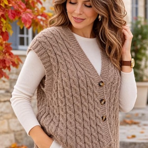 Peut inclure: Un gilet sans manches en tricot torsadé marron clair avec un col en V, des bordures côtelées et trois boutons marron foncé sur le devant. Le vêtement est porté sur un haut blanc à manches longues en maille épaisse et texturée.