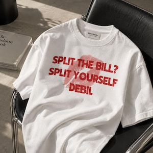 May include: A white short-sleeved t-shirt featuring red text that reads SPLIT THE BILL? SPLIT YOURSELF DEBIL, overlaid with a red lipstick kiss mark graphic. The shirt has a crew neckline and a branded label on the inside of the collar.