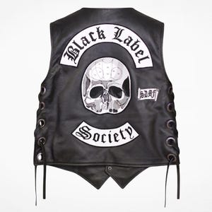 Puede incluir: Un chaleco de cuero negro con cordones laterales y ojales metálicos. La parte posterior muestra un parche blanco grande con un gráfico de calavera, el texto Black Label Society en fuente gótica y un parche rectangular pequeño con el texto SDMF.