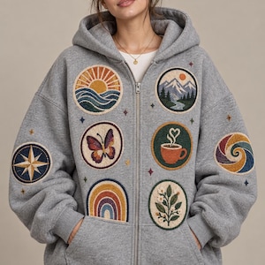 Puede incluir: Una sudadera con capucha y cremallera en color gris jaspeado que presenta múltiples parches circulares bordados. Los diseños incluyen un sol sobre olas, montañas, una mariposa, una taza de café, un arcoíris, una rosa de los vientos y flores.