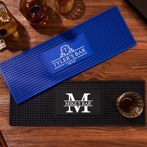 May include: Two rectangular rubber bar mats with a textured nub surface. One mat is blue with the white text "TYLER'S BAR" and "21st July 2026" inside a decorative frame. The other mat is black with a large white letter "M" and "MIKE'S BAR".