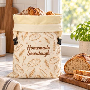 May include: A cream-colored fabric bread bag with a roll-top closure secured by black plastic buckles. The bag features a brown printed pattern of wheat stalks, rolling pins, spoons, and loaves, with the text Homemade Sourdough.
