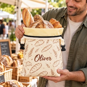 May include: A cream-colored fabric bread bag featuring a brown printed pattern of various loaves, croissants, and wheat stalks. The bag has a black zipper closure, black side buckles, and the name Oliver printed in brown script on the front.