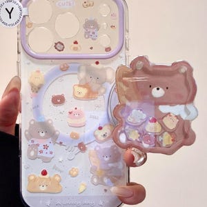 Puede incluir: Una funda de teléfono transparente con marco de cámara lavanda y un agarre en forma de oso. La funda presenta ilustraciones de osos, postres y el texto "CUTE!" y "A lovely day". El agarre de oso contiene pequeños dijes flotantes.