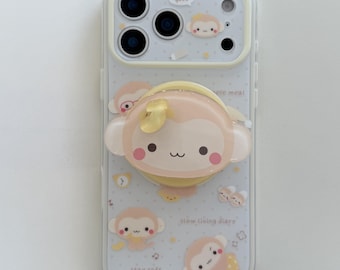 May include: A clear smartphone case featuring a light yellow camera frame and a central monkey-shaped pop-out grip. The case is decorated with small monkey illustrations, yellow dots, and the text "good," "simple meal," "slow living diary," and "stay soft."